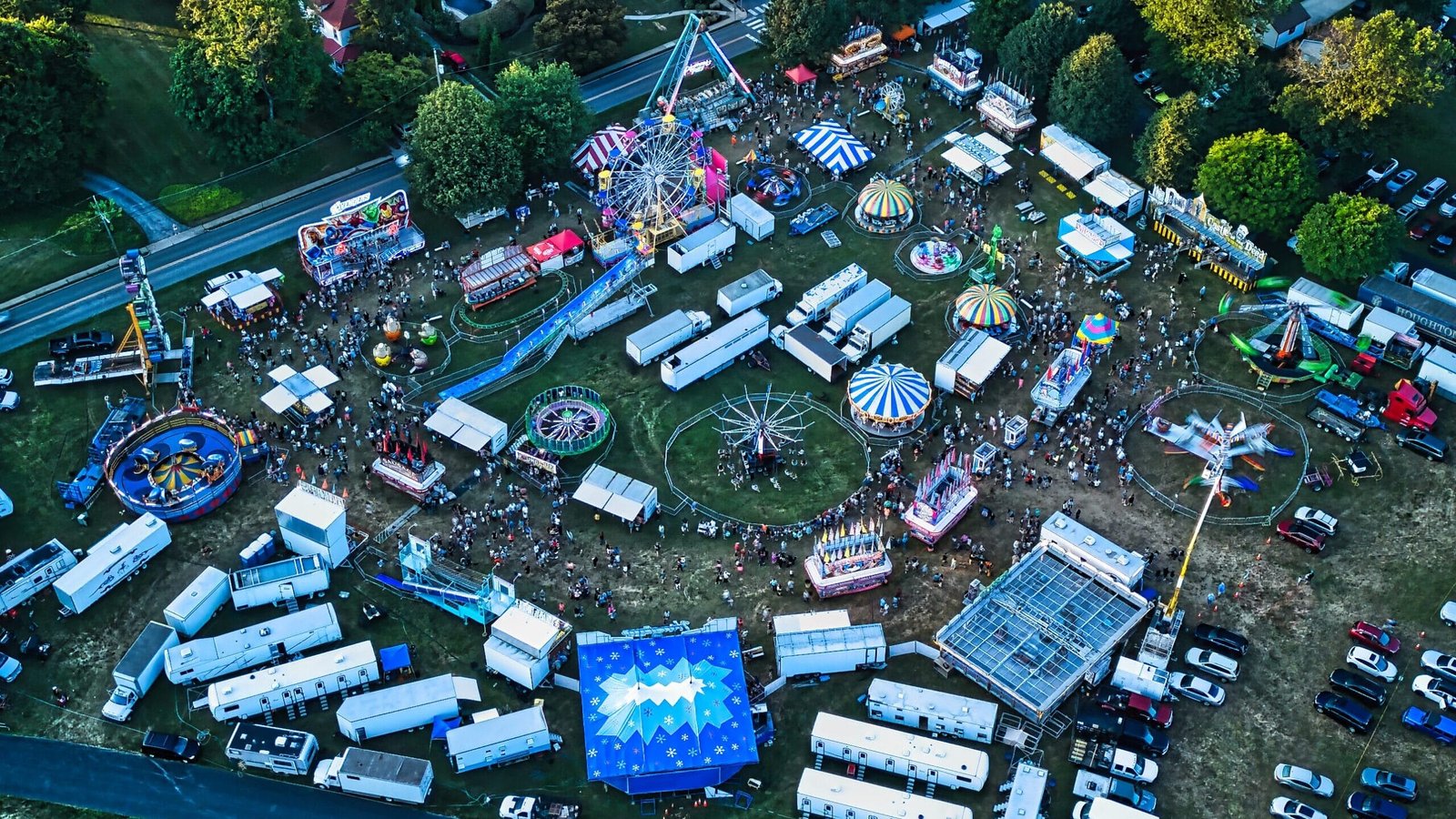 Aerial view of a carnival with rides, trailers, and vehicles on a grassy field next to a street and trees.