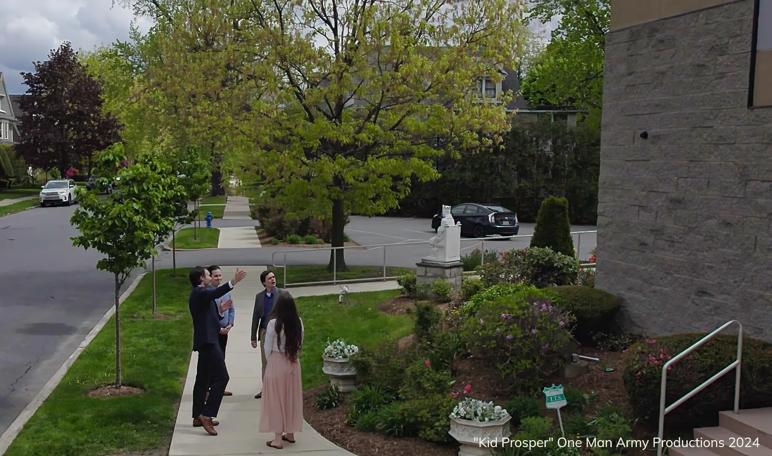 Four people standing on a sidewalk in front of a bulding with grass, a street, parking lot, vehicles, trees and houses in the background, and the text ""Kid Prosper" One Man Army Productions 2024" in the bottom right corner.