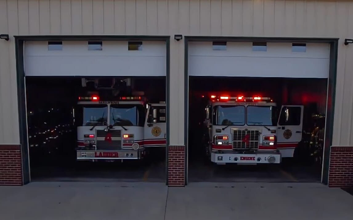 Building with two open garage doors and a white and red ladder firetruck inside the left bay and a red and white engine firetruck on the right side, both with red lights going on the top.