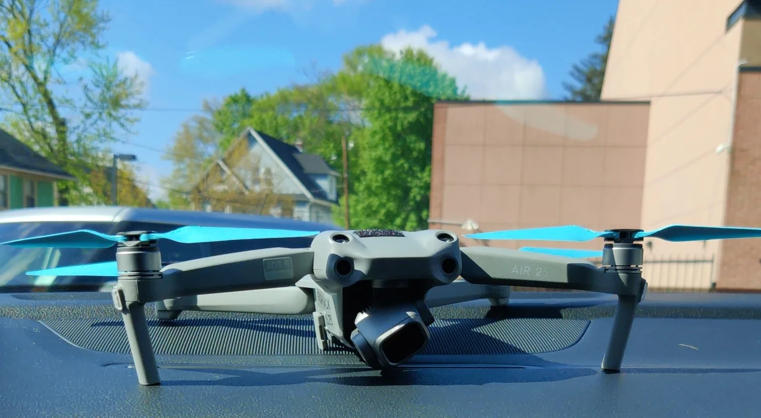 Gray drone with blue blades sitting on a black vehicle dashboard with houses, trees, and a building in the background.
